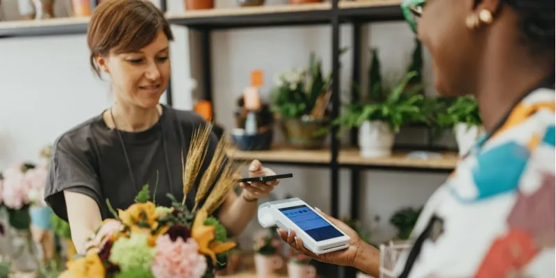 A customer is paying for a floral arrangement at a flower shop counter by tapping her smartphone to a point-of-sale device held by the cashier for a contactless transaction.